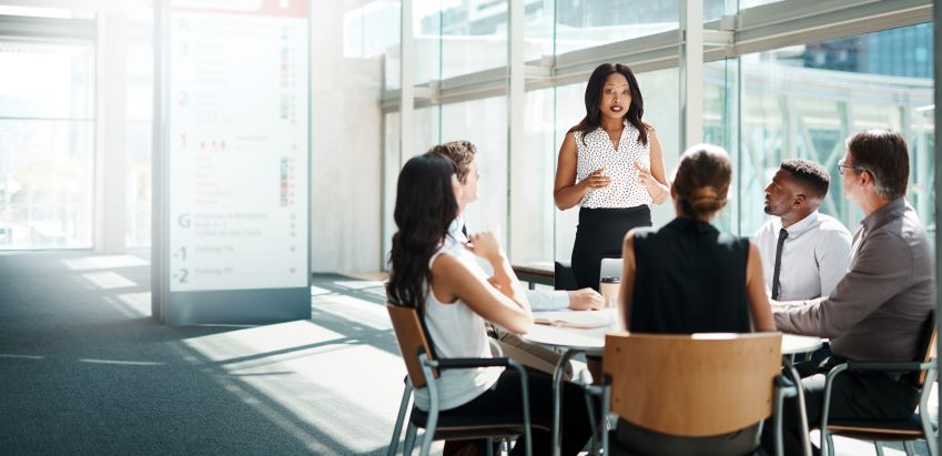 Alt - The image shows a group of five people seated around a table in a modern office setting, with one person standing and appearing to lead a discussion or presentation. The room has large windows allowing natural light to fill the space. There is a blurred signboard in the background with text and numbers. The scene appears to be a business meeting or a collaborative work session.