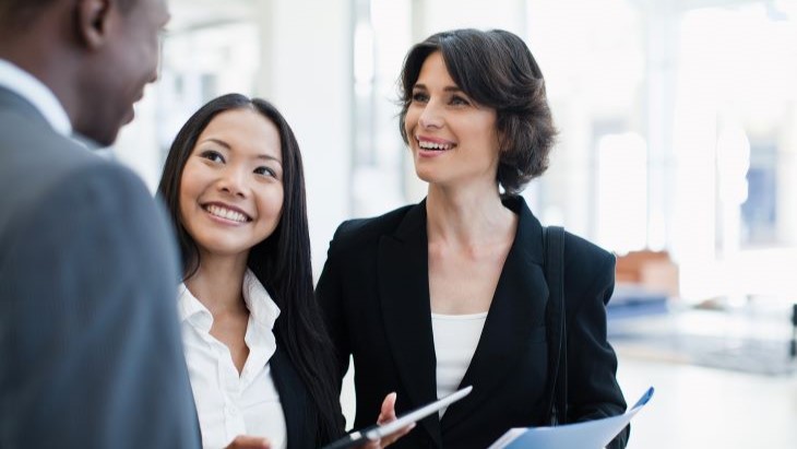 Alt - Three professionals engaged in a conversation in a bright, modern office setting. Two women, one with long black hair and the other with short wavy brown hair, are smiling while talking with a man in a suit. One woman holds a tablet, while the other has a folder in hand.