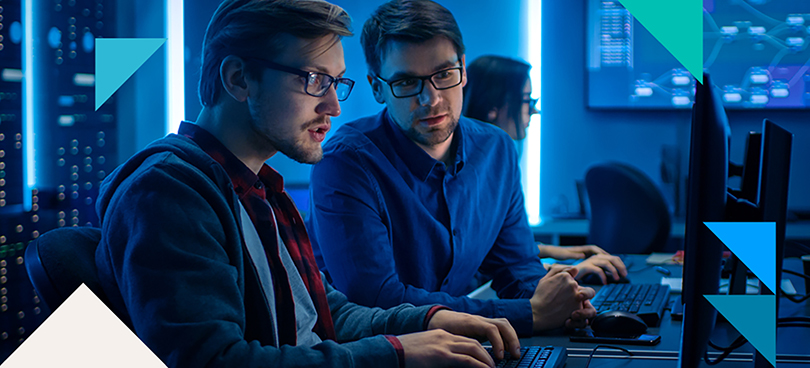 Two men in an office environment work on computers, one of them is leaning over to see the other man's screen.