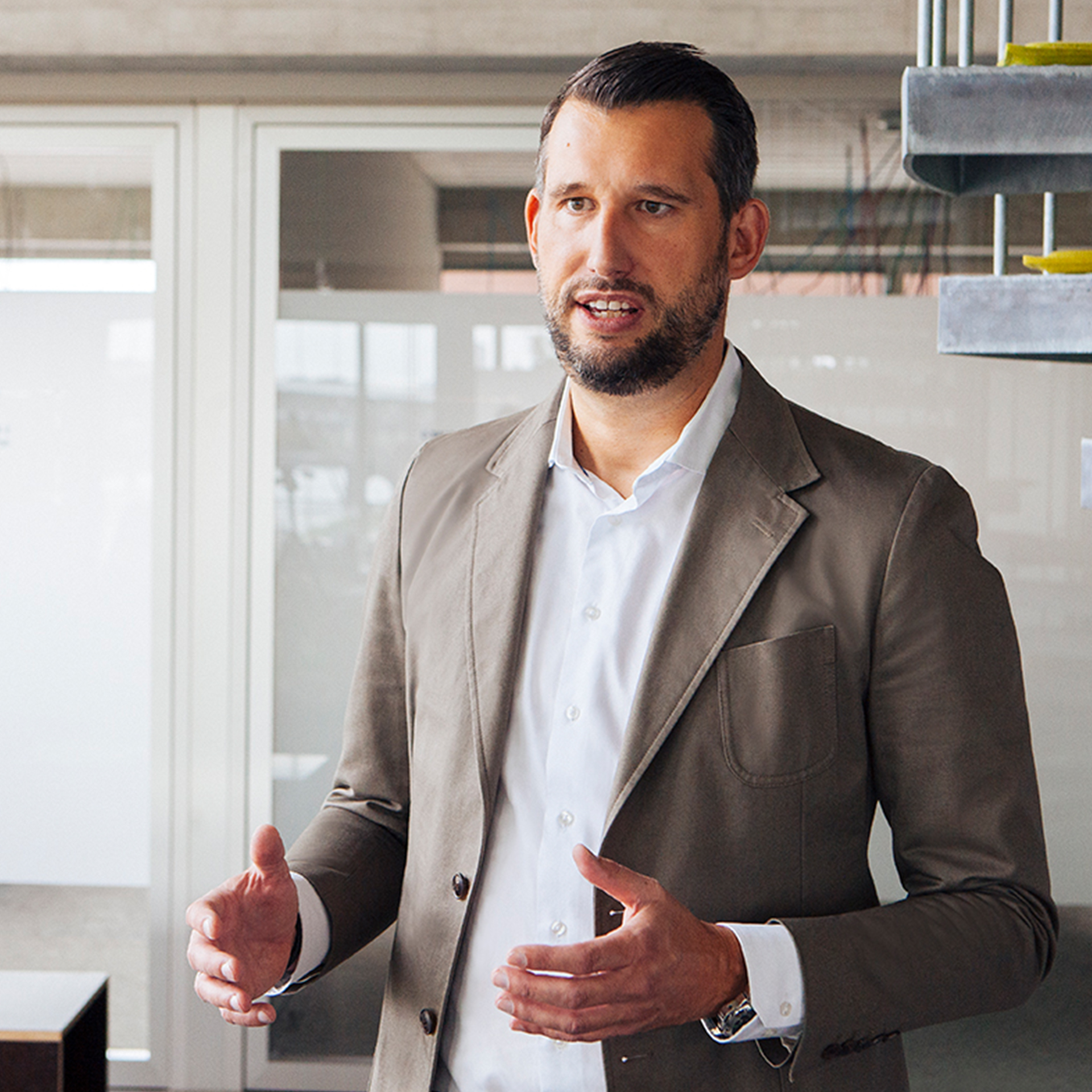 A smartly dressed man with a beard stands in front of a window. He is talking and gesturing with his hands.